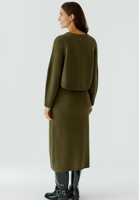 Gebreide jurk in olijfgroen met een cropped sweater overlay, lange mouwen en geribbelde boorden, gecombineerd met zwart glanzende laarzen.