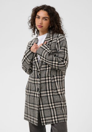 Femme aux cheveux bouclés portant un manteau à carreaux noir et blanc par-dessus une chemise blanche, tenant les revers du manteau avec les deux mains.