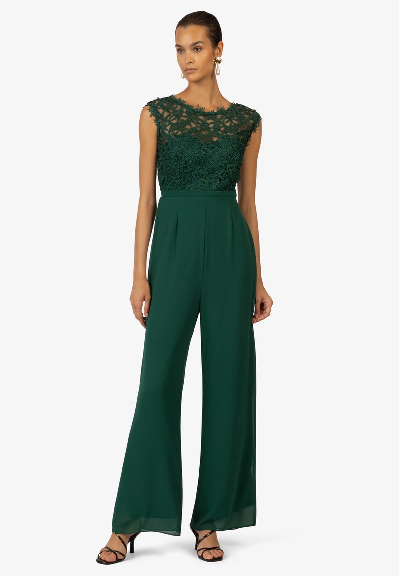 Green jumpsuit with a lace bodice featuring floral patterns, wide legs, and a fitted waist. Worn with strappy black heels.