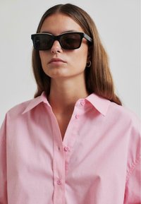 Camicia button-up rosa chiaro con colletto classico e bottoni rosa, abbinata a occhiali da sole neri rettangolari. Tessuto morbido, dettagli minimi.