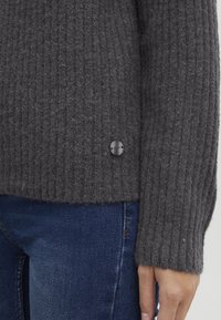 Primo piano di una persona che indossa un maglione a coste grigio scuro e jeans in denim blu, con un piccolo marchio metallico rotondo sul maglione.