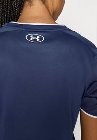 Tengerészkék sportpóló fehér díszítéssel, sima, légáteresztő anyagból készült. Hátul a nyak vonalánál kör alakú logó található.