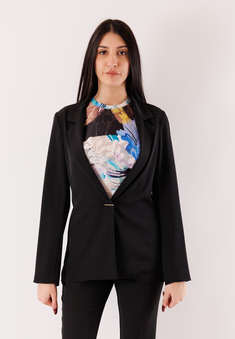 Blazer nero con colletto strutturato, chiusura con un singolo bottone dorato, indossato sopra una maglia a fantasia floreale colorata. Tessuto liscio, vestibilità sartoriale.