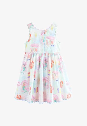 Lächelndes junges Mädchen in einem ärmellosen weißen Kleid mit bunten Motiven zum Thema Meer und passenden Sandalen vor neutralem Hintergrund.