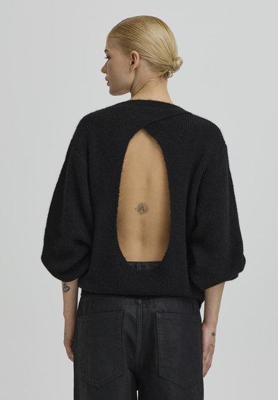 Szőke hajú nő kontyban, fekete pulóvert visel, amelynek hátulja nyitott, megmutatva a felső hátán lévő kis tetoválást.