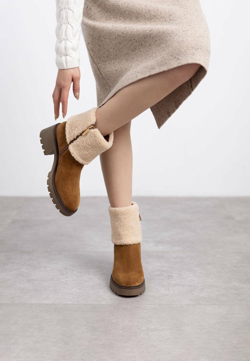 Stivale di suede marrone con un risvolto beige peloso e dettagli con zip. Suola piatta spessa con fondo testurizzato per una buona aderenza. Indossato con un abito a maglia beige.