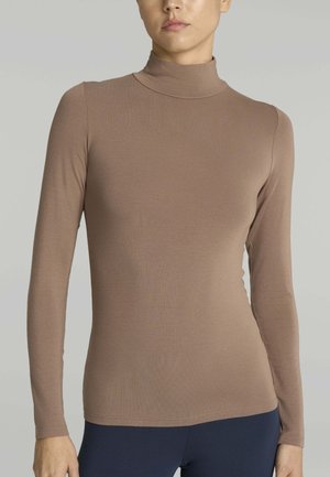 Persona che indossa un maglione a collo alto marrone aderente e pantaloni blu scuro, in piedi contro uno sfondo grigio chiaro semplice.