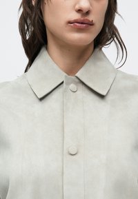 Hellgraue Wildlederjacke mit einem umgelegten Kragen, Knopfverschluss und glatter Textur, die ein sauberes, minimalistisches Design aufweist.