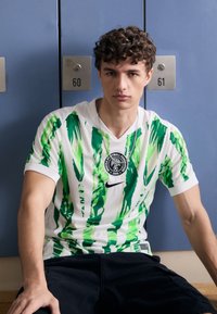 Chemise blanche à manches courtes avec de larges rayures vertes, dotée d'un badge circulaire sur la poitrine et d'un logo Nike. Un short noir complète la tenue.