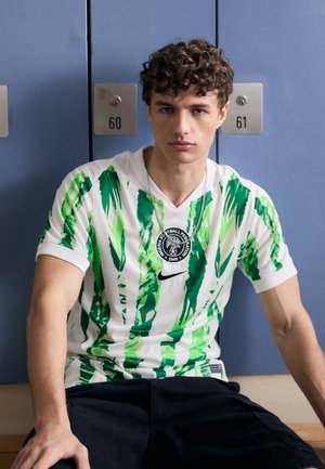 Camisa de manga corta blanca con rayas verdes llamativas, que presenta un distintivo circular en el pecho y un logo de Nike. Unos pantalones cortos negros completan el atuendo.