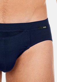 Slip da bagno blu navy in un tessuto morbido con una forma sagomata, caratterizzato da cuciture sottili e un piccolo logo nero sulla cintura.