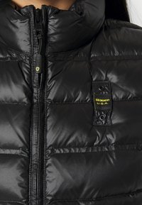 Czarna puchowa kamizelka z wysokim kołnierzem, zapinana z przodu na zamek, z wyrazistymi pikowaniami i żółtą metką "Blauer U.S.A." na piersi.