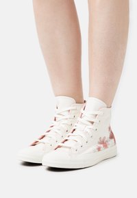 Bílé vysoké tenisky s květinovými růžovými akcenty, plátěný materiál a gumové podrážky. Obsahují tradiční šněrování a texturovaný povrch.