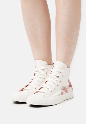 Bílé vysoké tenisky s květinovými růžovými akcenty, plátěný materiál a gumové podrážky. Obsahují tradiční šněrování a texturovaný povrch.
