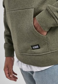 Olivgrüner Kapuzenpulli mit Fronttasche, gerippten Bündchen und Saum, ausgestattet mit einem kleinen schwarzen Etikett mit weißem Text am unteren Rand.