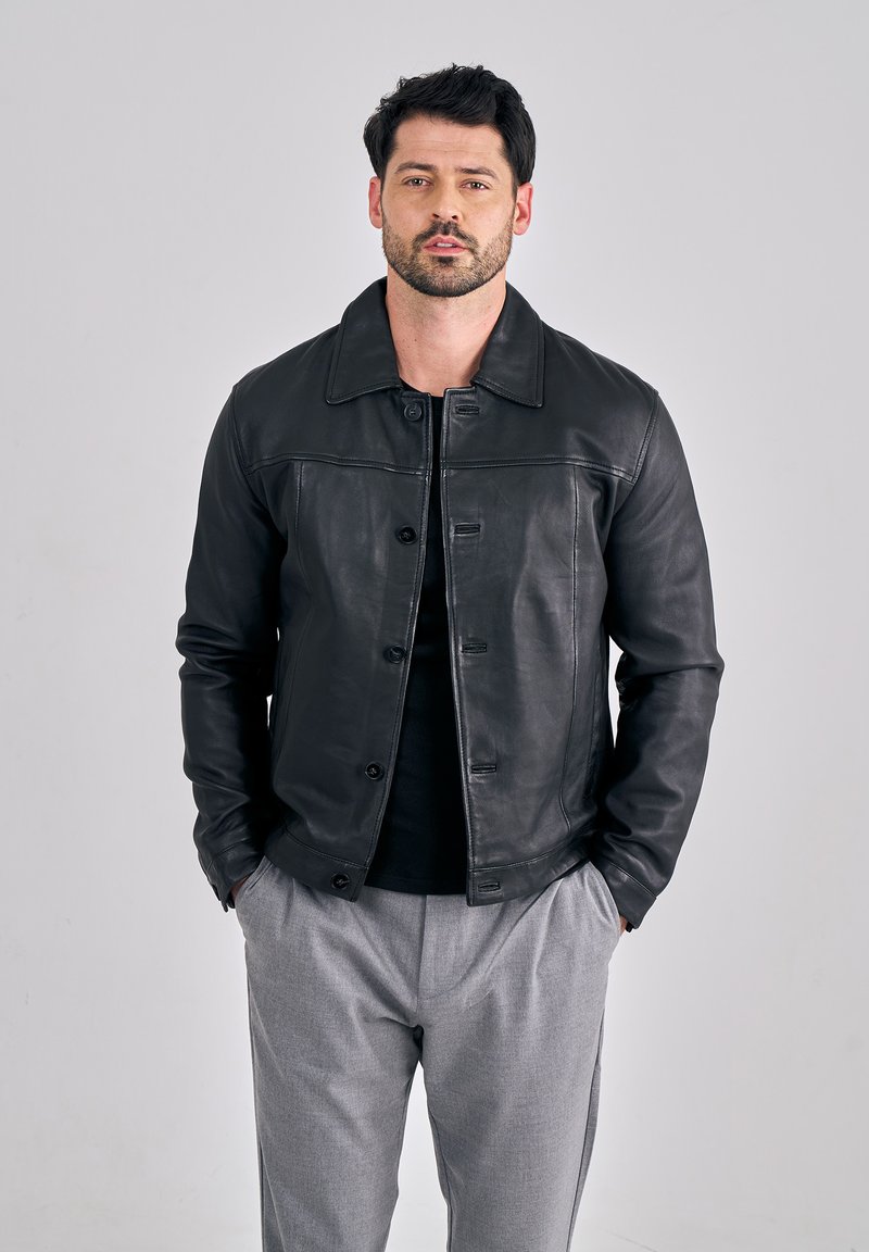 Schwarze Lederjacke mit Kragen, Vorderknöpfen und strukturierter Oberfläche, getragen über einem dunklen Oberteil und grauen Hosen.