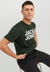 Camiseta de algodón verde con texto en blanco audaz que dice "JACK & JONES". Manga corta y cuello redondo. Modelo sentado en una silla.
