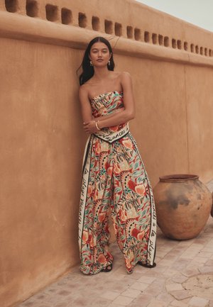 Femme portant une combinaison fleurie bustier à jambes larges avec des détails manuscrits, debout contre un mur texturé en terre cuite à côté d'un grand pot en argile.