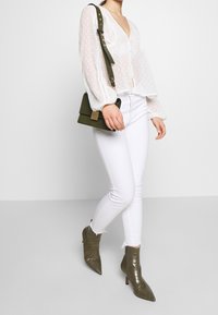 Weiße Bluse mit Tupfenmuster und Puffärmeln, kombiniert mit weißen Skinny-Jeans und olivgrünen Stiefeletten mit Absatz. Kleine Umhängetasche mit Riemen.