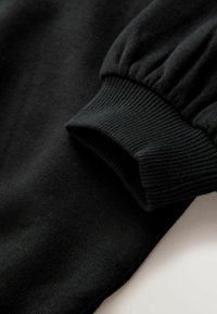 Schwarzes Langarmkleidungsstück mit geripptem elastischem Bündchen, nahaufgenommen auf einer hellen Oberfläche.