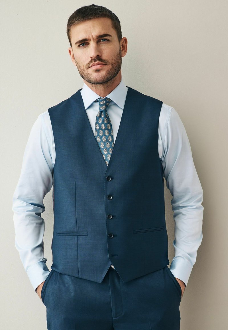 Gilet blu navy con finitura testurizzata, dotato di cinque bottoni e tasche laterali, indossato sopra una camicia azzurra chiara con una cravatta a fantasia.