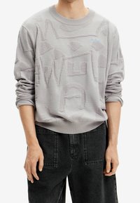 Jasnoszary sweter o kroju crewneck z teksturowanymi abstrakcyjnymi wzorami. Posiada prążkowane mankiety i dół, zestawiony z ciemnymi spodniami dżinsowymi.