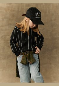 Svart strikket genser med hvite vertikale striper, lyseblå jeans, og en grønn rutete flanellskjorte knyttet rundt livet. Svart caps med brodert tekst.