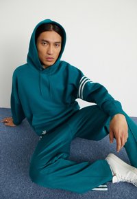 Felpa teal con cappuccio regolabile e tasca frontale, caratterizzata da tre strisce bianche sul braccio sinistro. Abbinata a pantaloni jogger teal e scarpe da ginnastica bianche.
