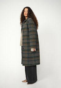 Karierten Wollmantel in grünen, braunen und grauen Tönen, mit kragenförmigem Design, Knopfverschluss und seitlichen Taschen. Getragen über einem beigen Pullover.