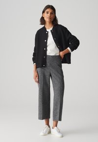 Zwarte gebreide bomberjack met drukknoopsluiting over een witte blouse, gecombineerd met grijze geweven brede broek en witte sneakers.