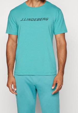 Mężczyzna w turkusowej koszuli z krótkim rękawem z napisem „J.LINDEBERG” oraz pasujących spodniach, stojący z luźno opuszczonymi ramionami.