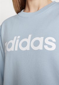 Osoba v světle modré mikině s velkým bílým logem "adidas" přes hrudník, viditelná od krku po horní část trupu.