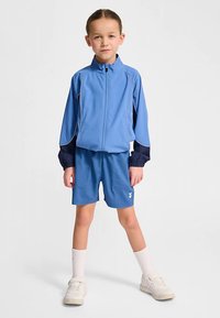 Blå sportjacka med marinblå detaljer, matchad med blå shorts. Har lättviktstyg, dragkedja och logotypdetalj på shortsen. Vita sneakers och strumpor.