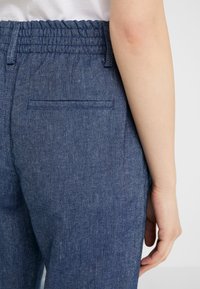 Närbild av en persons midja och arm som bär blå byxor med elastisk midja och en springa bakficka, mot en enkel ljus bakgrund.