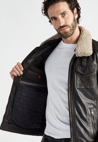 Man met krullend haar en baard die een donker leren jasje met schapenvachtkraag opent, waarbij de zwarte binnenvoering en een wit T-shirt eronder zichtbaar worden.