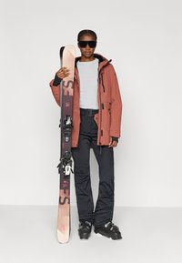 Σετ ρούχων για σκι με αδιάβροχο μπουφάν σε σκουριά χρώμα, γκρι πουκάμισο, μαύρο παντελόνι και μαύρες μπότες. Κρατώντας μαύρα και ροζ πέδιλα με δεσίματα.