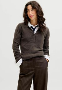 Žena s valovitom tamnom kosom, koja nosi smeđi pulover preko bijele košulje i tamnih hlača s prugicama, s rukama u džepovima, blago se okreće desno.