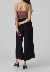 Kvinna med lockigt hår bär en mauvevirkad crop top, svarta vida byxor, svarta klacksandaler och ett guldarmband, står vänd bort.
