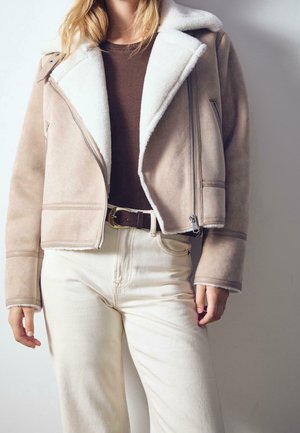 Woman wearing beige shearling jacket over brown top, cream high-waist pants, and dark belt against a plain background.