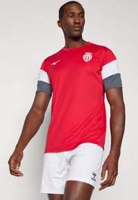 Camiseta deportiva roja de manga corta con acentos en blanco y gris, que presenta un logo en el pecho. Se lleva con pantalones cortos blancos. Textura suave.