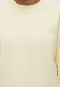 Personne portant un sweat-shirt uni jaune clair à col rond, montrant la partie supérieure de la poitrine et du cou.
