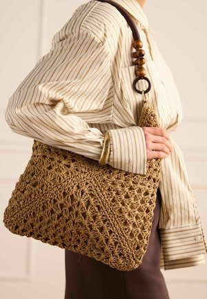 Femme portant une chemise beige à rayures et une jupe foncée, tenant un grand sac à bandoulière marron en crochet avec une sangle à perles en bois.