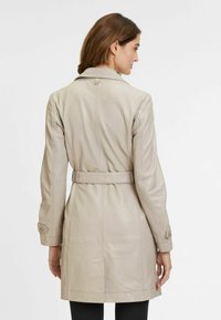 Cappotto in pelle beige con vita cintrata, maniche lunghe e polsini con bottoni, caratterizzato da una texture liscia e un design su misura.
