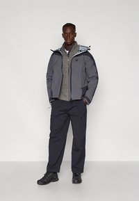 Giacca imbottita grigia con cappuccio, chiusura frontale con zip e pantaloni grigio scuro. Maglione strutturato sotto, calzature nere. Design semplice, stile funzionale.