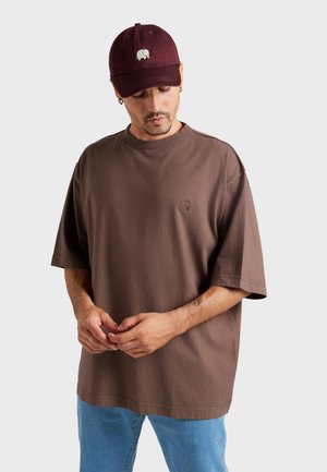 Homme portant un T-shirt marron oversize et une casquette de baseball bordeaux avec un logo d’éléphant, debout devant un fond clair uni.