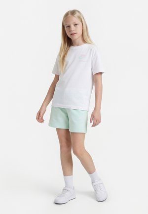 Enfant blond debout, portant un t-shirt blanc Nike, un short Nike vert clair, des chaussettes blanches et des baskets blanches, sur fond blanc uni.