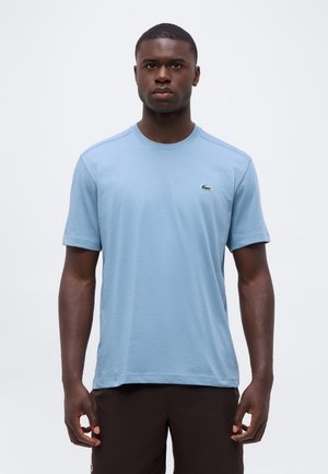 Junger Mann trägt ein schlichtes hellblaues T-Shirt mit kurzen Ärmeln und einem kleinen grünen Krokodil-Logo auf der linken Brust sowie dunkle Hosen.