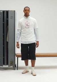 Wit T-shirt met Arsenal-logo, over een blauw gestreept overhemd gedragen. Zwarte knielange korte broek, witte sokken en Adidas-sneakers met rode strepen.