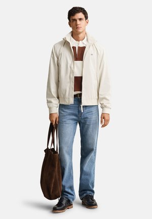 Young man wearing a beige zip jacket, brown and white striped polo, blue jeans, black shoes, holding a large brown tote bag in his left hand.