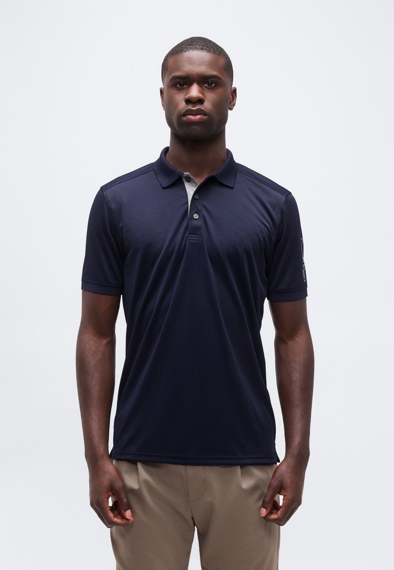 Άνδρας που στέκεται μπροστά σε λευκό φόντο, φορά μπλε σκούρο πόλο πουκάμισο και μπεζ παντελόνι, με ουδέτερη έκφραση.